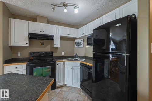 Kitchen with black appliances, dark countertops, a textured ceiling, white cabinetry, and rail lighting - 314 5340 199 Street, Edmonton, AB - Indoor Photo Showing Kitchen With Double Sink