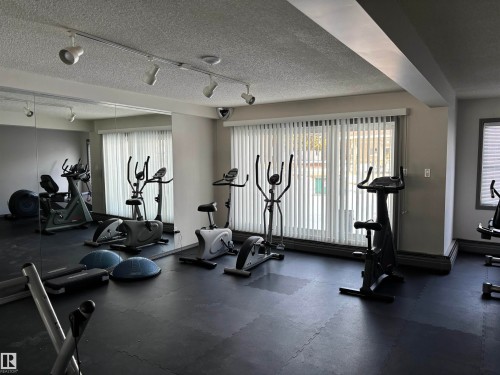 Dedicated fitness room featuring interlocking rubber floor tiles, full-wall mirror, track lighting, and vertical blinds on a large window - 207 11465 41 Avenue, Edmonton, AB - Indoor Photo Showing Gym Room