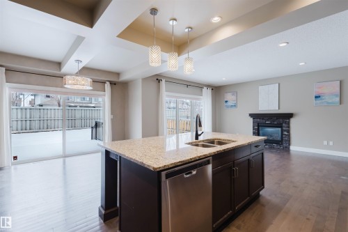 Kitchen with open floor plan, stainless steel dishwasher, dark wood finished floors, pendant lighting, and light stone counters - 1957 Ainslie Link, Edmonton, AB - Indoor Photo Showing Kitchen With Fireplace With Double Sink