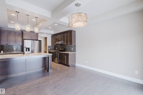 Kitchen featuring dark wood finish cabinets, stainless steel appliances, coffered ceiling, light stone countertops, and decorative backsplash - 1957 Ainslie Link, Edmonton, AB - Indoor Photo Showing Kitchen With Upgraded Kitchen