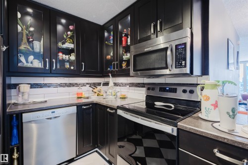 Kitchen featuring dark cabinets, stainless steel appliances, glass fronted cabinets, backsplash, and a textured ceiling - 1803 10149 Saskatchewan Drive, Edmonton, AB - Indoor Photo Showing Kitchen