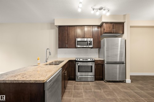 Kitchen featuring dark wood finish cabinetry, stainless steel appliances, light stone counters, and a peninsula - 29 11518 76 Avenue, Edmonton, AB - Indoor Photo Showing Kitchen With Stainless Steel Kitchen With Double Sink With Upgraded Kitchen
