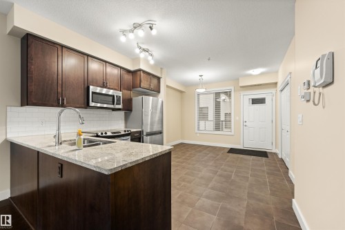 Kitchen with dark wood finish cabinets, a peninsula, stainless steel appliances, tasteful backsplash, and light stone counters - 29 11518 76 Avenue, Edmonton, AB - Indoor Photo Showing Kitchen With Stainless Steel Kitchen With Double Sink With Upgraded Kitchen