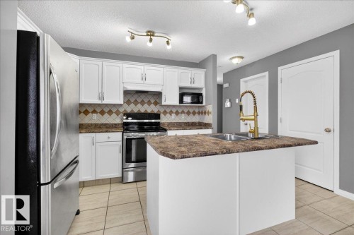 Kitchen with stainless steel appliances, dark countertops, white cabinets, a center island with sink, and light tile patterned flooring - 150 Acacia Circle, Leduc, AB - Indoor Photo Showing Kitchen With Double Sink