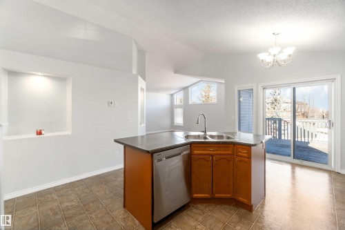 Kitchen featuring dark countertops, a kitchen island with sink, stainless steel dishwasher, wood finish cabinets, and vaulted ceiling - 20604 48 Avenue, Edmonton, AB - Indoor Photo Showing Kitchen With Double Sink