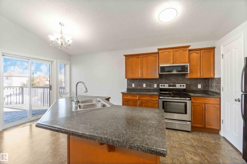 Kitchen featuring stainless steel appliances, dark countertops, a kitchen island with sink, decorative backsplash, and wood finish cabinets - 20604 48 Avenue, Edmonton, AB - Indoor Photo Showing Kitchen With Double Sink