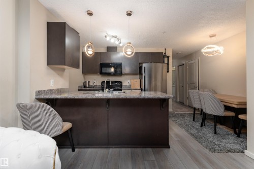 Kitchen with a peninsula, dark wood finish cabinetry, black appliances, a kitchen breakfast bar, and a textured ceiling - 205 340 Windermere Road, Edmonton, AB - Indoor Photo Showing Kitchen With Upgraded Kitchen