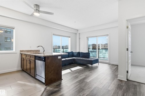 Kitchen featuring a peninsula, light stone counters, light wood finished floors, stainless steel dishwasher, and a textured ceiling - 106 1238 Windermere Way, Edmonton, AB - Indoor