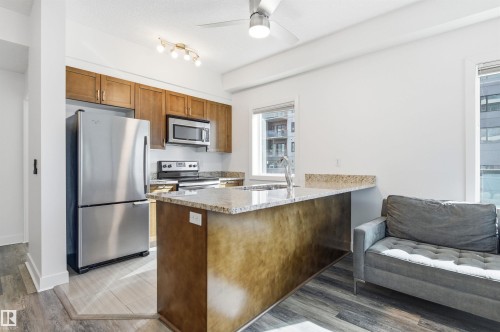 Kitchen featuring stainless steel appliances, a peninsula, wood finish cabinets, ceiling fan, and light stone countertops - 106 1238 Windermere Way, Edmonton, AB - Indoor Photo Showing Kitchen