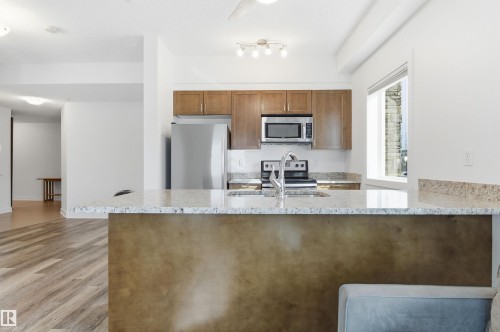 Kitchen featuring a peninsula, light stone countertops, stainless steel appliances, wood finish cabinetry, and light wood finished floors - 106 1238 Windermere Way, Edmonton, AB - Indoor Photo Showing Kitchen With Double Sink