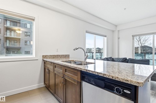 Kitchen with a peninsula, light stone counters, stainless steel dishwasher, open floor plan, and wood finish cabinets - 106 1238 Windermere Way, Edmonton, AB - Indoor Photo Showing Kitchen With Double Sink