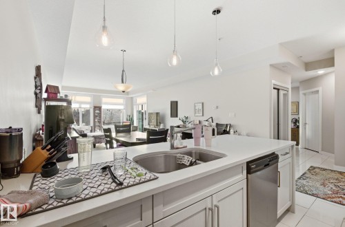 Kitchen featuring dishwasher, open floor plan, white cabinetry, and light stone countertops - 309 625 Leger Way, Edmonton, AB - Indoor Photo Showing Kitchen With Double Sink