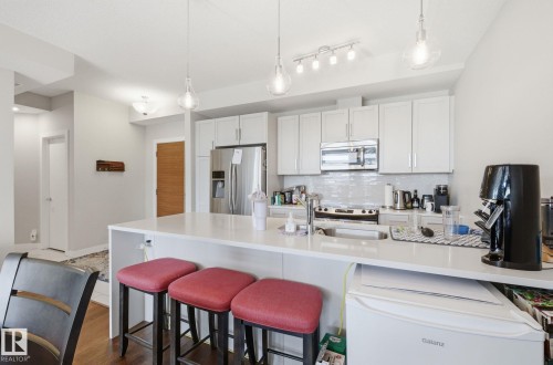 Kitchen featuring a breakfast bar, stainless steel appliances, white cabinetry, decorative light fixtures, and decorative backsplash - 309 625 Leger Way, Edmonton, AB - Indoor Photo Showing Kitchen