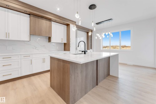 Two tone kitchen with two tone cabinets, light stone counters, a center island with sink, light wood-type flooring, and decorative backsplash - 13 Trill Point(E), Spruce Grove, AB - Indoor Photo Showing Kitchen With Upgraded Kitchen