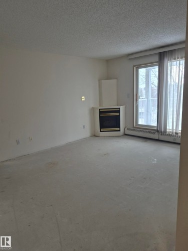 Unfurnished living room with a glass covered fireplace, baseboard heating, a textured ceiling, and unfinished concrete flooring - 410 17467 98A Avenue, Edmonton, AB - Indoor With Fireplace