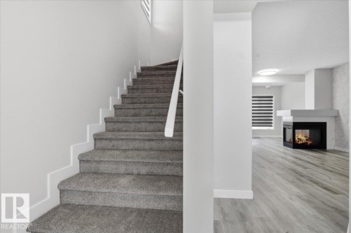 The property features a staircase with carpeted steps and a white railing, alongside a living area with light-colored flooring, a modern fireplace, and a window with horizontal blinds - 6 6410 134 Avenue Nw, Edmonton, AB - Indoor Photo Showing Other Room With Fireplace