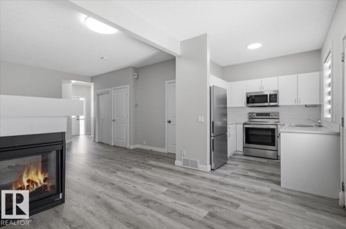 Open concept living area featuring a fireplace, grey wood-look flooring, and a kitchen with white cabinetry and stainless steel appliances - 6 6410 134 Avenue Nw, Edmonton, AB - Indoor Photo Showing Other Room With Fireplace