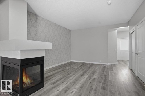 The living area features a modern fireplace, light-toned wood-look flooring, and an accent wall with patterned wallpaper - 6 6410 134 Avenue Nw, Edmonton, AB - Indoor With Fireplace
