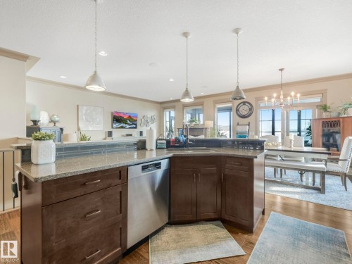 Kitchen featuring dark wood finish cabinetry, stainless steel dishwasher, light stone counters, hanging lights, and open floor plan - 117 Mcdowell Wynd, Leduc, AB - Indoor Photo Showing Kitchen With Upgraded Kitchen