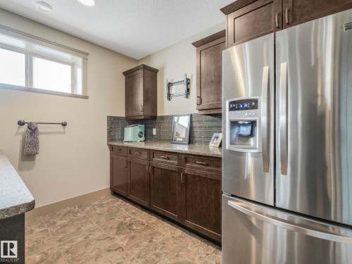 Kitchen with stainless steel fridge with ice dispenser, decorative backsplash, dark wood finish cabinetry, a textured ceiling, and light stone counters - 117 Mcdowell Wynd, Leduc, AB - Indoor Photo Showing Kitchen