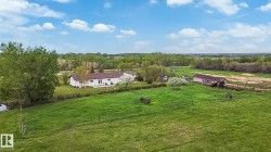Expansive green pasture surrounding a white ranch-style home with dark roofing, mature trees, and an outbuilding - 