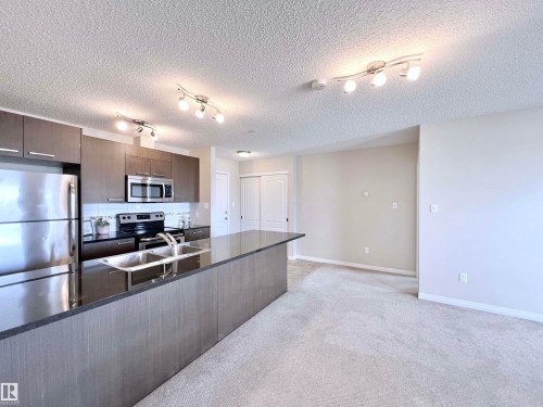 Kitchen featuring stainless steel appliances, dark wood finish cabinets, track lighting, light colored carpet, and backsplash - 3308 9351 Simpson Drive, Edmonton, AB - Indoor Photo Showing Kitchen With Double Sink