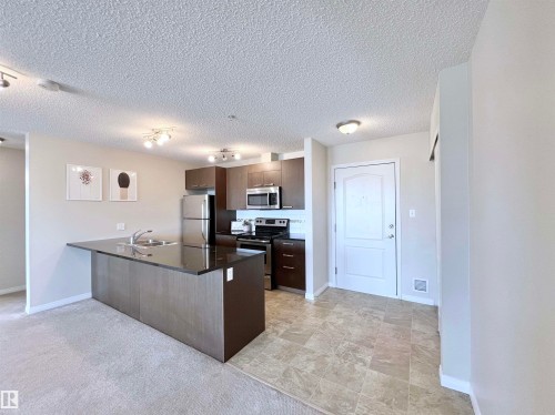 Kitchen featuring a peninsula, stainless steel appliances, light colored carpet, a textured ceiling, and dark wood finish cabinetry - 3308 9351 Simpson Drive, Edmonton, AB - Indoor Photo Showing Kitchen