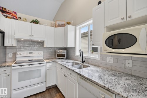 Kitchen with light stone countertops, white appliances, white cabinets, vaulted ceiling, and dark wood-style floors - 30 4 Heritage Way, St. Albert, AB - Indoor Photo Showing Kitchen With Double Sink