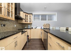 Kitchen with light wood finish cabinetry, stainless steel dishwasher, dark wood finished floors, dark stone counters, and glass fronted cabinets - 