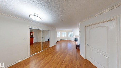 Unfurnished living room featuring light wood-style flooring, a glass covered fireplace, a textured ceiling, crown molding, and baseboard heating - 115 9938 104 St, Edmonton, AB - Indoor Photo Showing Other Room