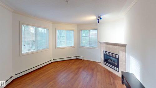 Unfurnished living room featuring a baseboard heating unit, a tiled fireplace, hardwood / wood-style flooring, and ornamental molding - 115 9938 104 St, Edmonton, AB - Indoor With Fireplace