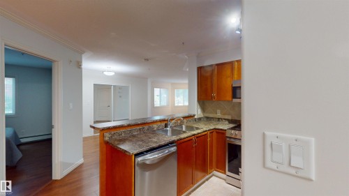 Kitchen with a peninsula, stainless steel appliances, wood finish cabinetry, dark countertops, and baseboard heating - 115 9938 104 St, Edmonton, AB - Indoor Photo Showing Kitchen With Double Sink