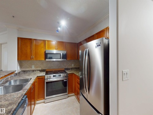 Kitchen with wood finish cabinetry, stainless steel appliances, backsplash, crown molding, and dark stone counters - 115 9938 104 St, Edmonton, AB - Indoor Photo Showing Kitchen With Double Sink