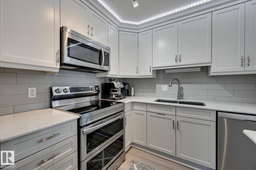 Kitchen featuring stainless steel appliances, light stone counters, light wood-type flooring, tasteful backsplash, and a textured ceiling - 59 14803 Miller Boulevard, Edmonton, AB - Indoor Photo Showing Kitchen With Double Sink