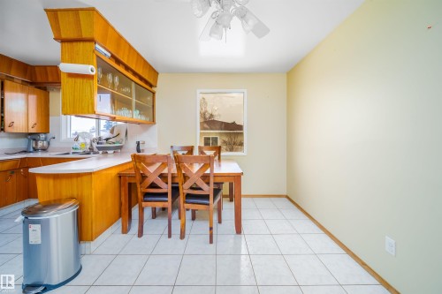 Kitchen and dining area featuring light-colored tile flooring, a window, and wooden cabinetry with glass-fronted upper cabinets - 7911 145 Avenue Nw, Edmonton, AB - Indoor