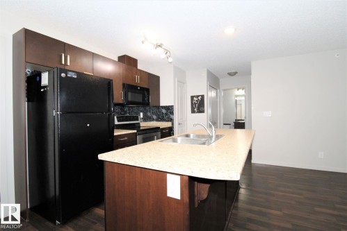 Kitchen with black appliances, a kitchen island with sink, dark wood finish cabinetry, light countertops, and dark wood-style floors - 430 5515 7 Avenue, Edmonton, AB - Indoor Photo Showing Kitchen With Double Sink