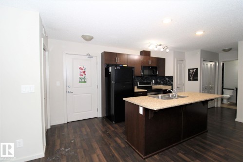 Kitchen featuring dark wood finish cabinetry, black appliances, a kitchen island with sink, a breakfast bar, and dark wood-type flooring - 430 5515 7 Avenue, Edmonton, AB - Indoor Photo Showing Kitchen With Double Sink