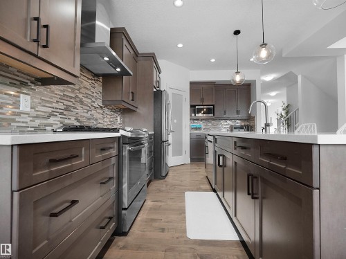 Kitchen featuring stainless steel appliances, dark wood finish cabinets, pendant lighting, light wood-style floors, and light stone counters - 3387 Parker Loop, Edmonton, AB - Indoor Photo Showing Kitchen With Upgraded Kitchen