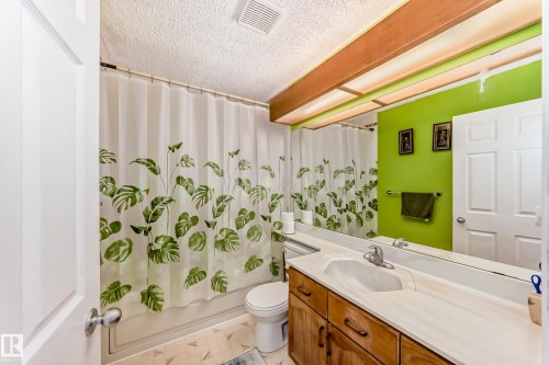 Bathroom featuring a wooden vanity with a light-colored countertop and an integrated sink, a mirror spanning the length of the vanity, and a shower with a white and green leaf-patterned curtain - 11532 9 Avenue, Edmonton, AB - Indoor Photo Showing Bathroom