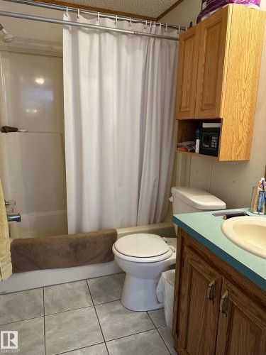The bathroom features a shower-tub combination with a shower curtain, a vanity with a sink and a light blue countertop, and upper wooden cabinetry - 67 4204 47 Street, Wetaskiwin, AB - Indoor Photo Showing Bathroom