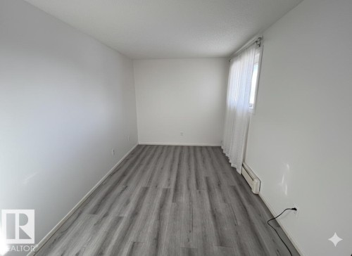This room features gray wood-style flooring, white walls, and a window with white sheer curtains - 212 2620 Mill Woods Road E, Edmonton, AB - Indoor Photo Showing Other Room