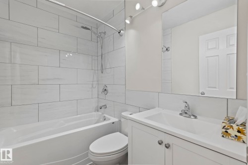 The bathroom features a bathtub with a shower, white tiled walls, a toilet, and a vanity with a sink and mirror - 18124 68Ave, Edmonton, AB - Indoor Photo Showing Bathroom