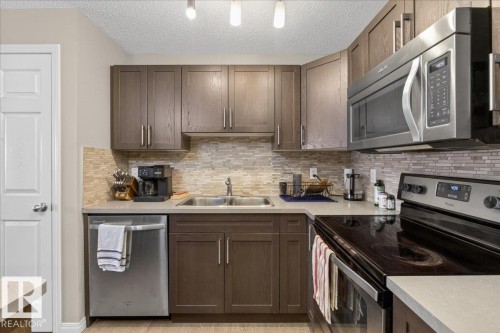 The kitchen features dark wood cabinetry, a tiled backsplash, and stainless steel appliances including a dishwasher, microwave, and stove - 33 4029 Orchards Drive Sw, Edmonton, AB - Indoor Photo Showing Kitchen With Double Sink