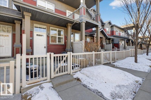 The property features a red and gray exterior with white trim, a white front door, and a white picket fence - 33 4029 Orchards Drive Sw, Edmonton, AB - Outdoor With Deck Patio Veranda With Facade