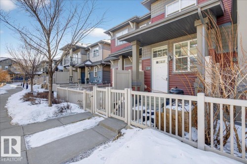 The property features a private fenced patio area, a white front door with an overhead transom window, and red siding on the exterior - 33 4029 Orchards Drive Sw, Edmonton, AB - Outdoor