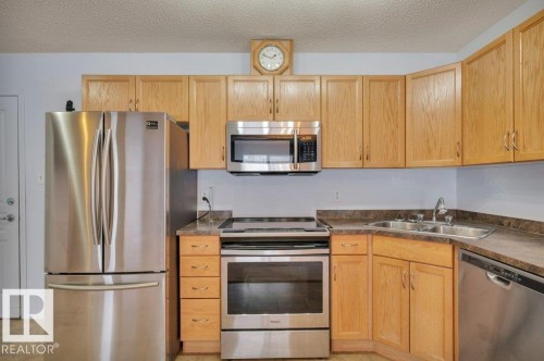 The kitchen features light wood cabinetry, stainless steel appliances including a refrigerator, oven, microwave, and dishwasher, and a double basin sink - 103 309 Clareview Station Drive, Edmonton, AB - Indoor Photo Showing Kitchen With Double Sink