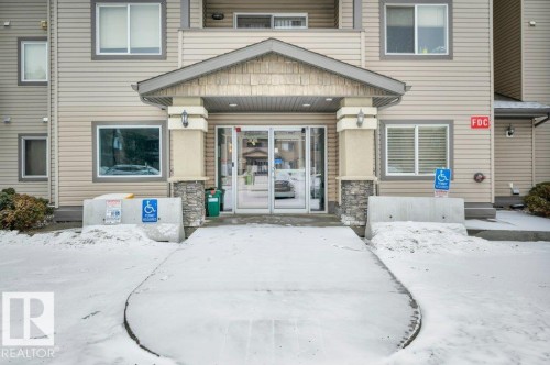 The property features a building exterior with light-colored siding, a covered entrance with stone accent pillars, and large glass doors - 103 309 Clareview Station Drive, Edmonton, AB - Outdoor With Facade