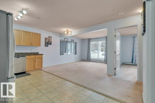 The property features an open-concept living area with light-colored carpeting, a kitchen area with tiled flooring, light wood cabinetry, and a stainless steel dishwasher - 103 309 Clareview Station Drive, Edmonton, AB - Indoor