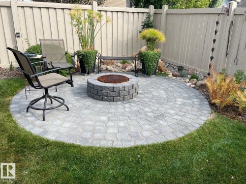 Outdoor living space featuring a circular paver patio with a stone fire pit, surrounded by lush green lawn and garden beds - 11 Etoile Cres N, St. Albert, AB - Outdoor