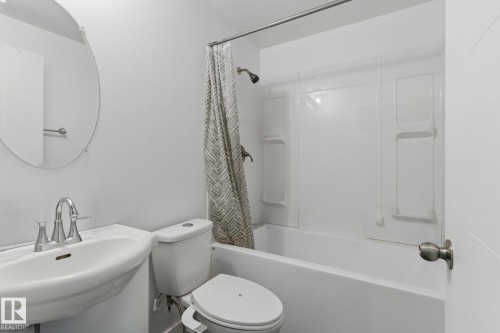 Bathroom featuring a pedestal sink with a chrome faucet, a round wall mirror, and a bathtub with a shower and a patterned shower curtain - 2416 Wonnacott Crest, Edmonton, AB - Indoor Photo Showing Bathroom